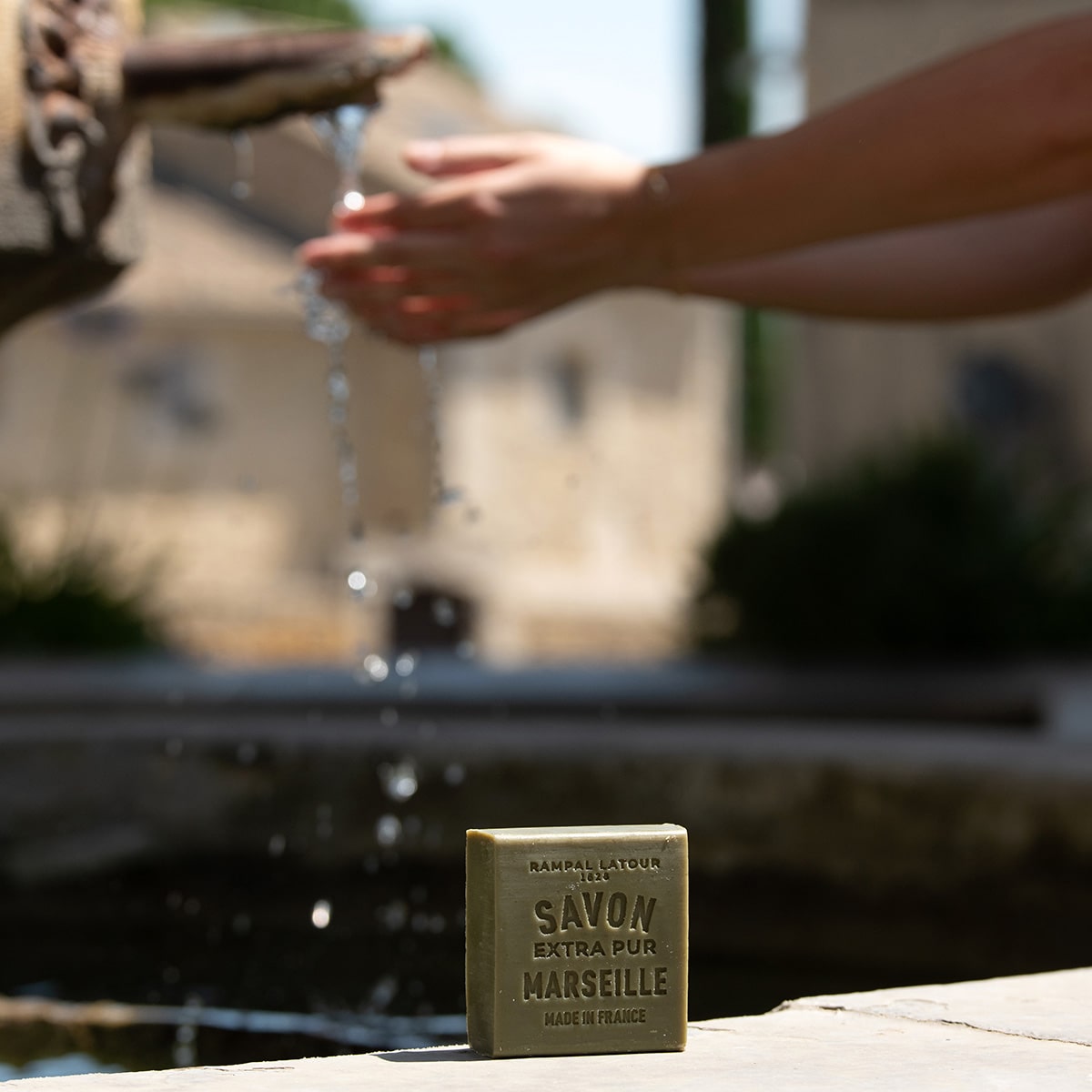 Savon de Marseille Rampal Latour posé au bord d'une fontaine, mains sous un filet d'eau
