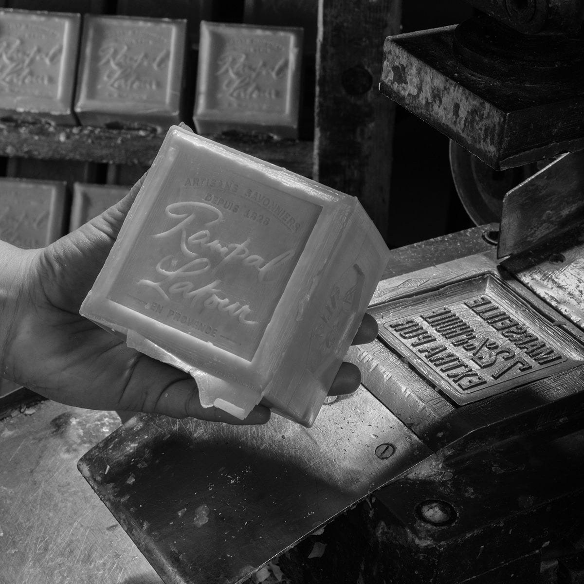 Cube de savon de Marseille Rampal Latour tenu à la main devant une presse de savonnerie traditionnelle